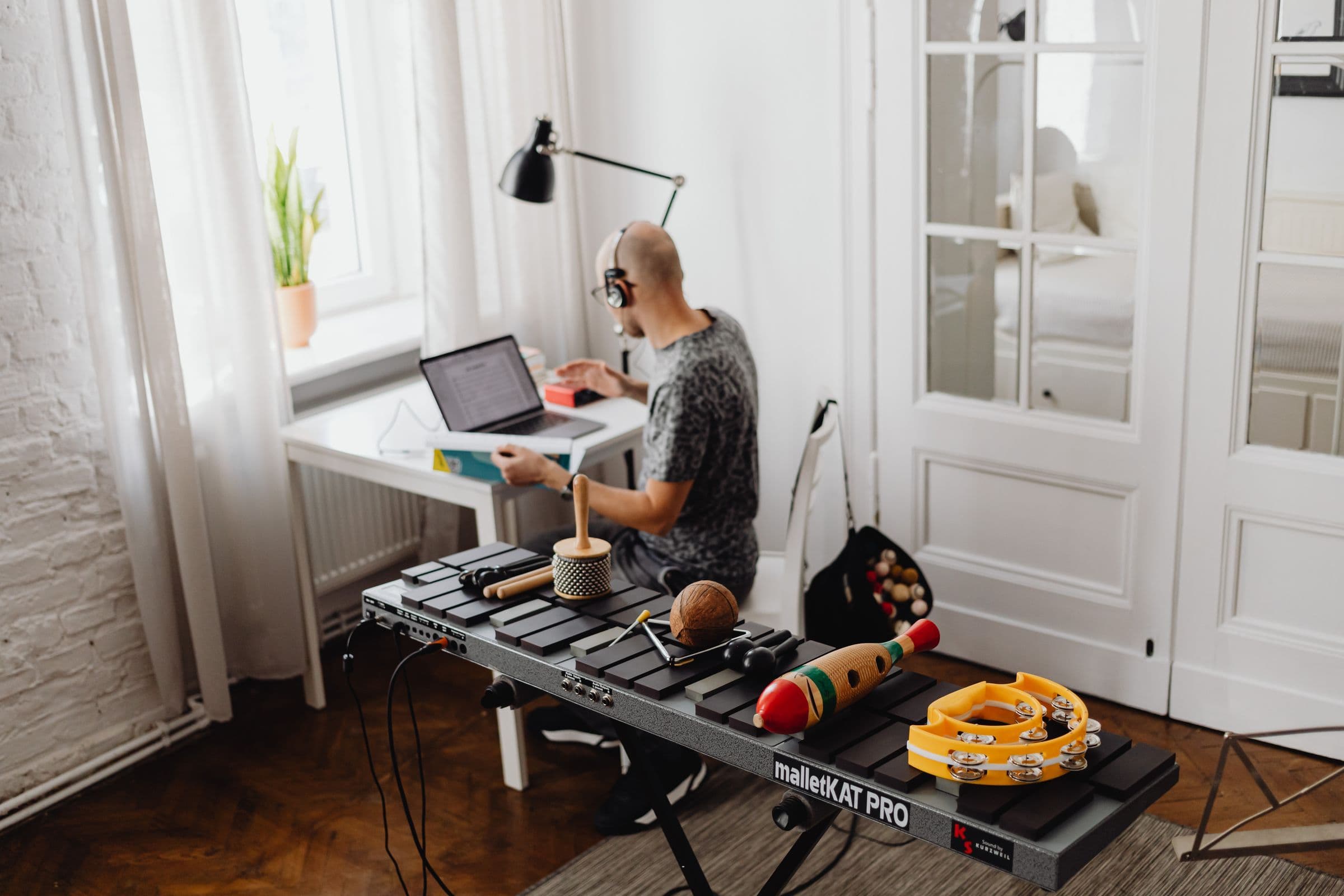 Musician at a laptop in a bright home studio with a xylophone and percussion