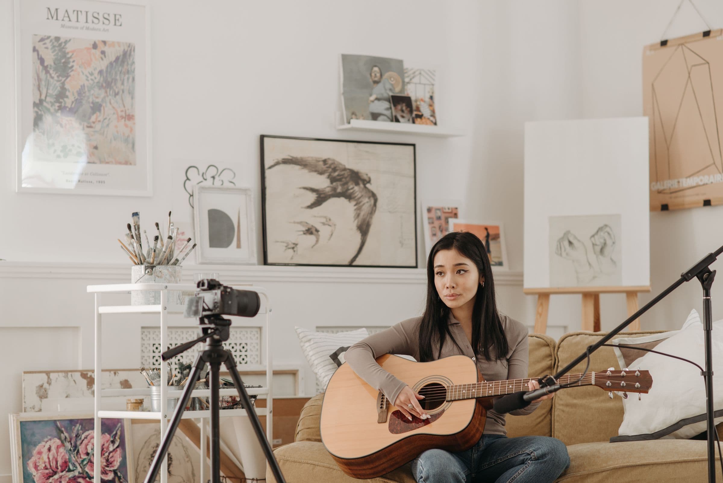 Musician playing acoustic guitar on a sofa while filming herself with a camera on a tripod
