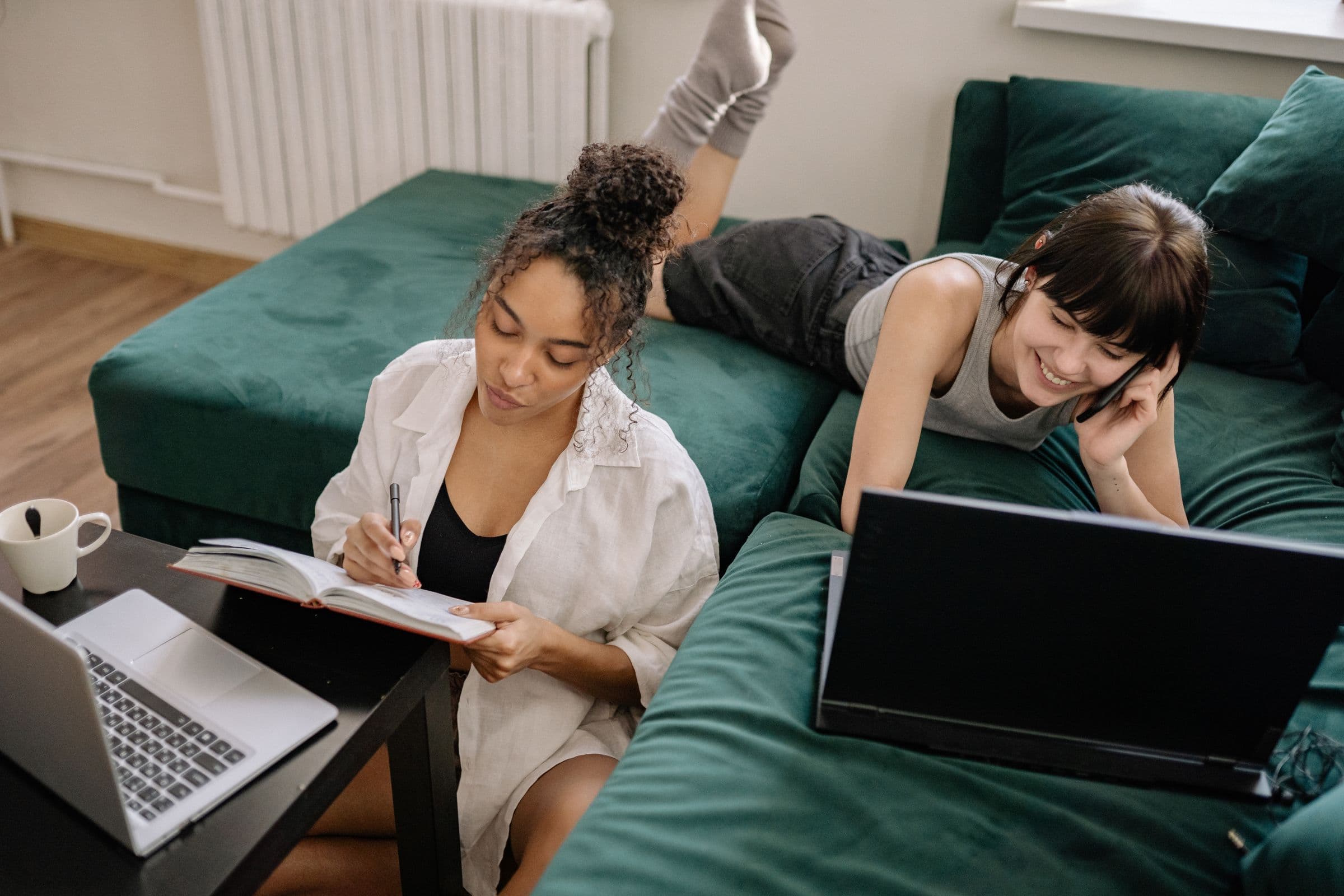 Two friends on a green sofa with laptops, one taking notes in a notebook