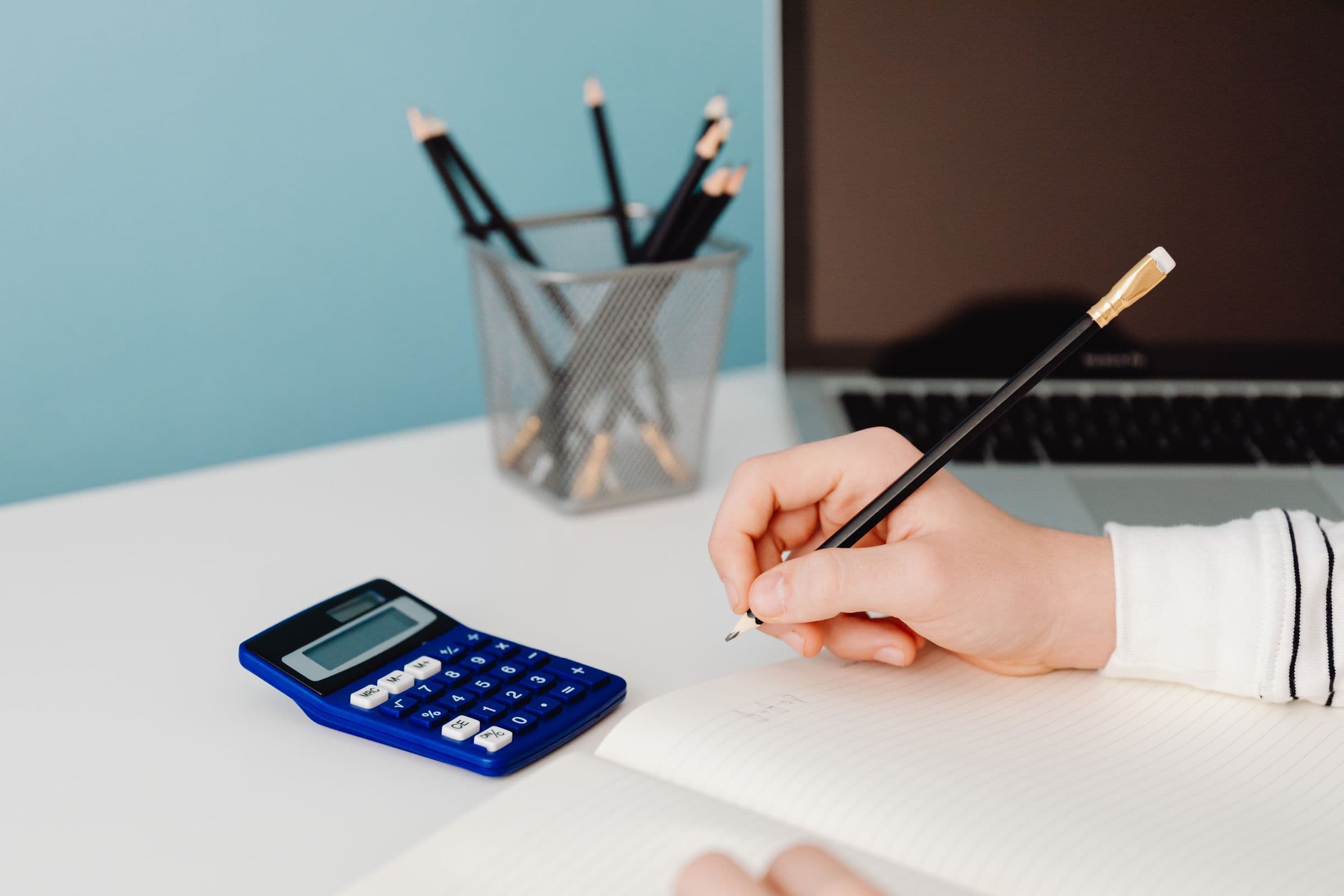 Hand writing in a notebook next to a blue calculator and a laptop