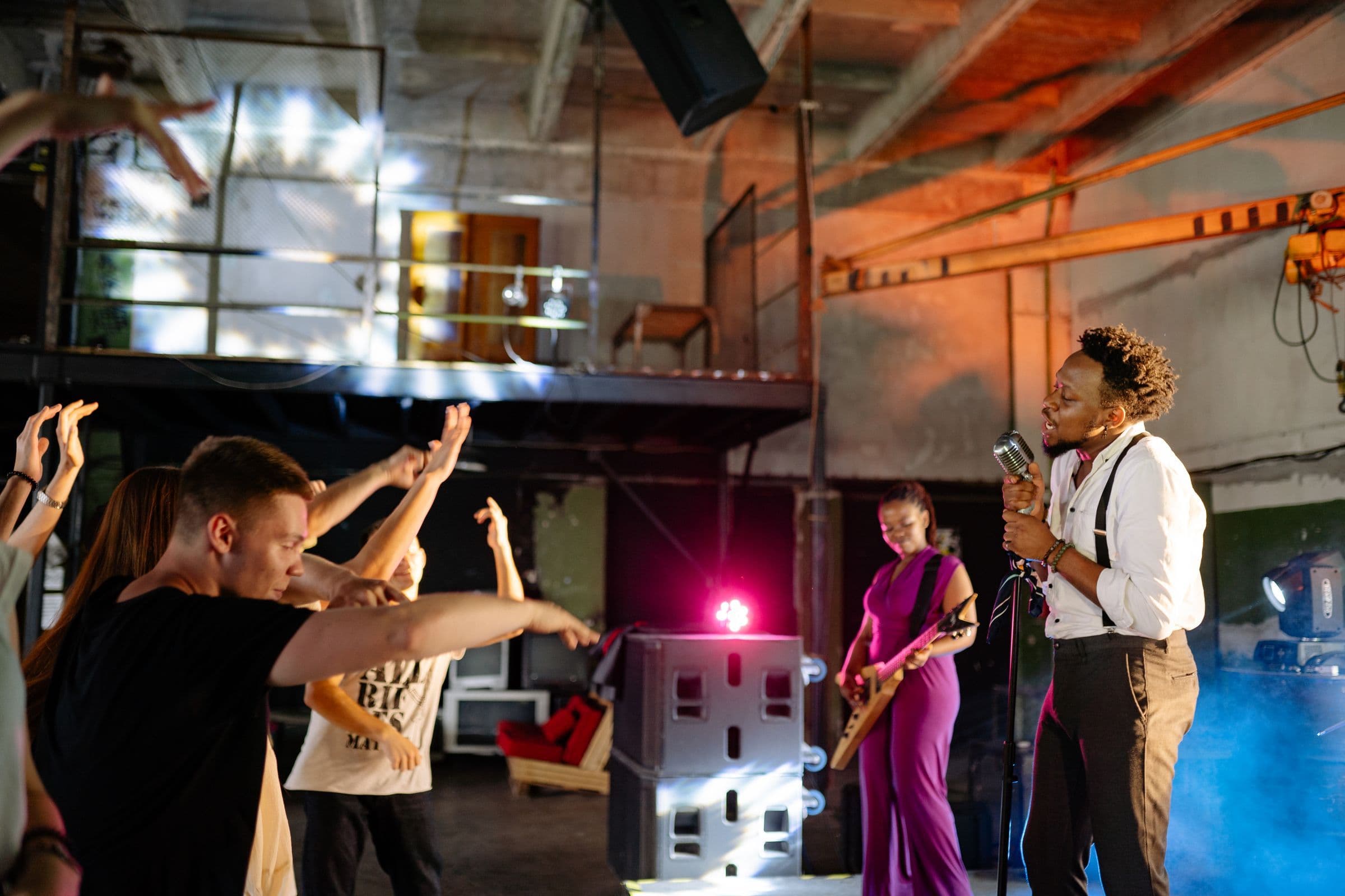 Singer in suspenders performing at a small industrial venue while the crowd raises their hands