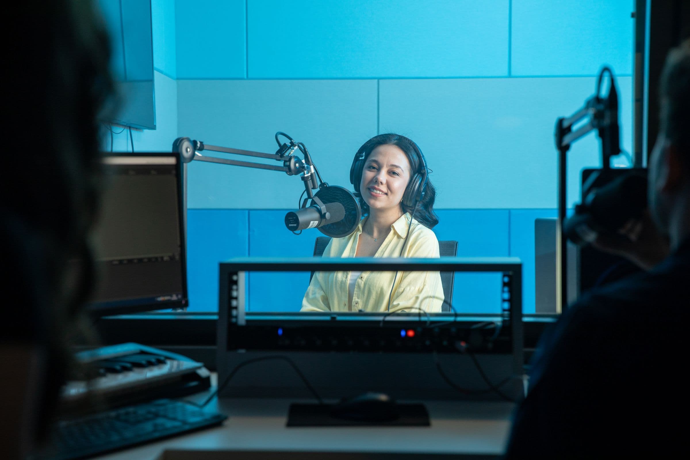 Host wearing headphones at a studio microphone, seen past the mixing desk