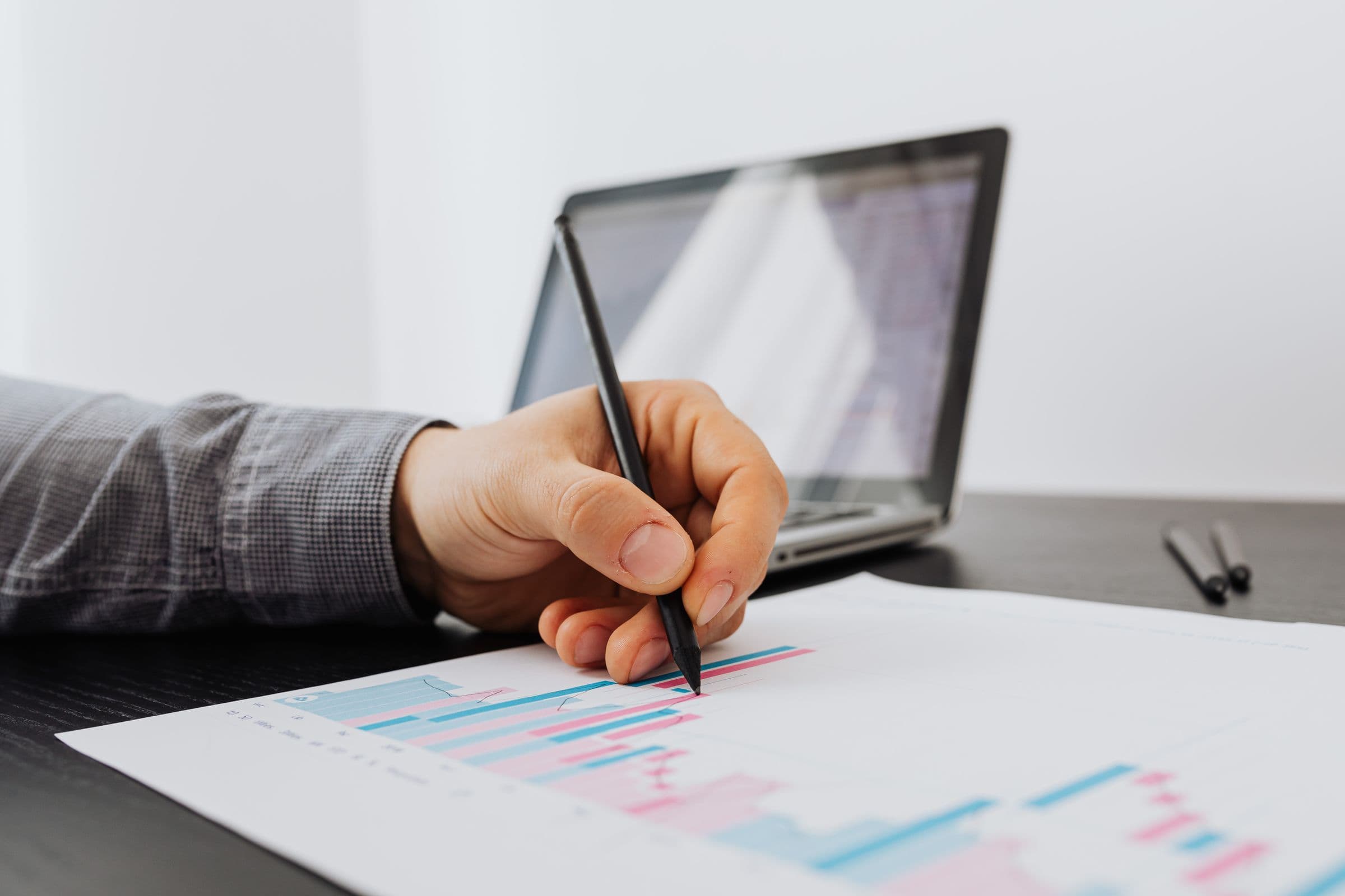 Hand with a pen marking a printed bar chart, with a laptop in the background