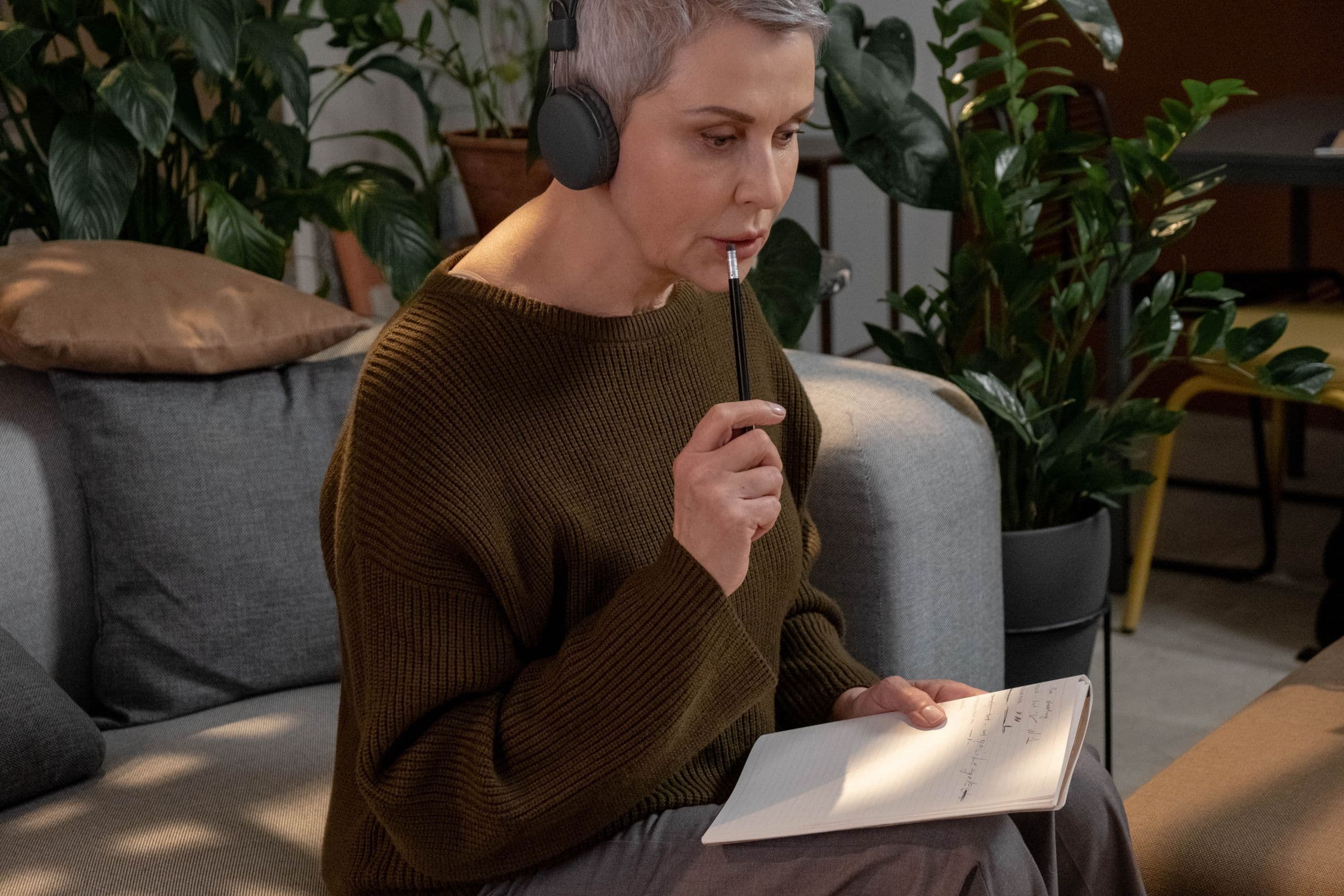 Woman in headphones on a couch, thinking with a pencil and a notebook