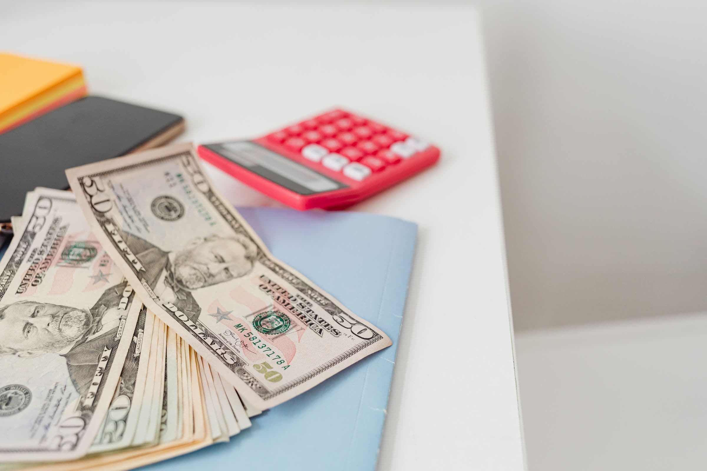 Fanned-out dollar bills next to a pink calculator on a white desk