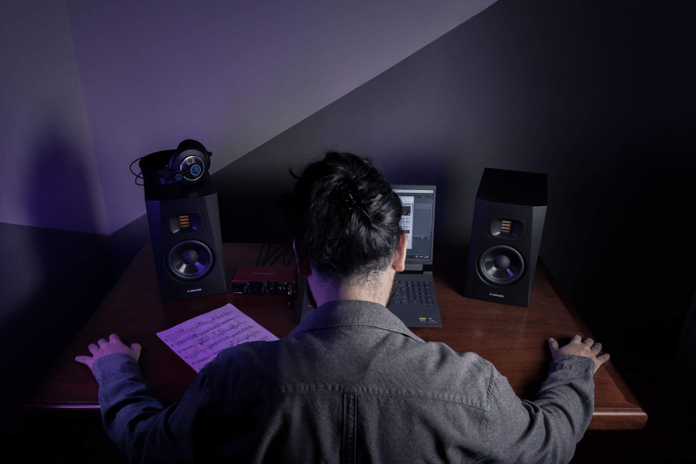 Musician at a home studio desk between two speakers, with a laptop and sheet music