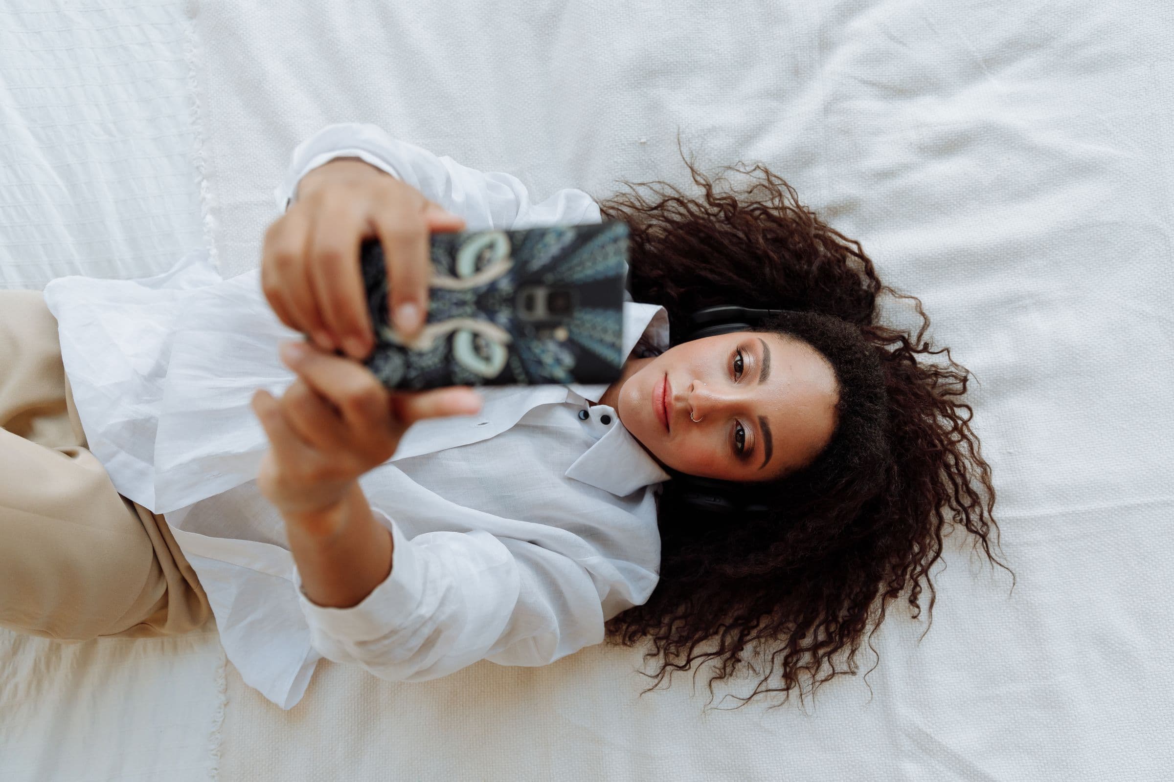 Woman in headphones lying on a white bed, looking at her phone