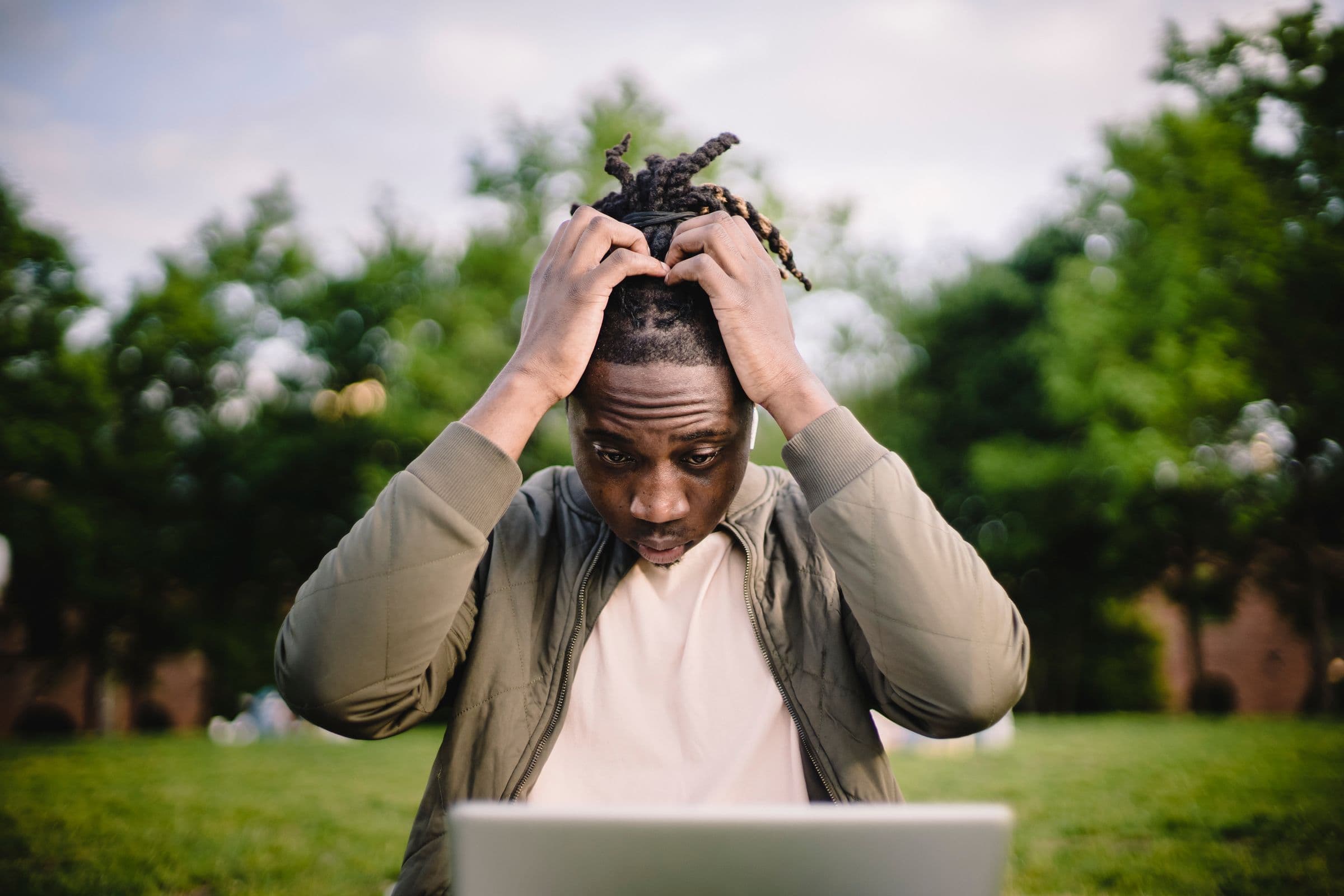 Young man holding his head in both hands while looking at a laptop outdoors