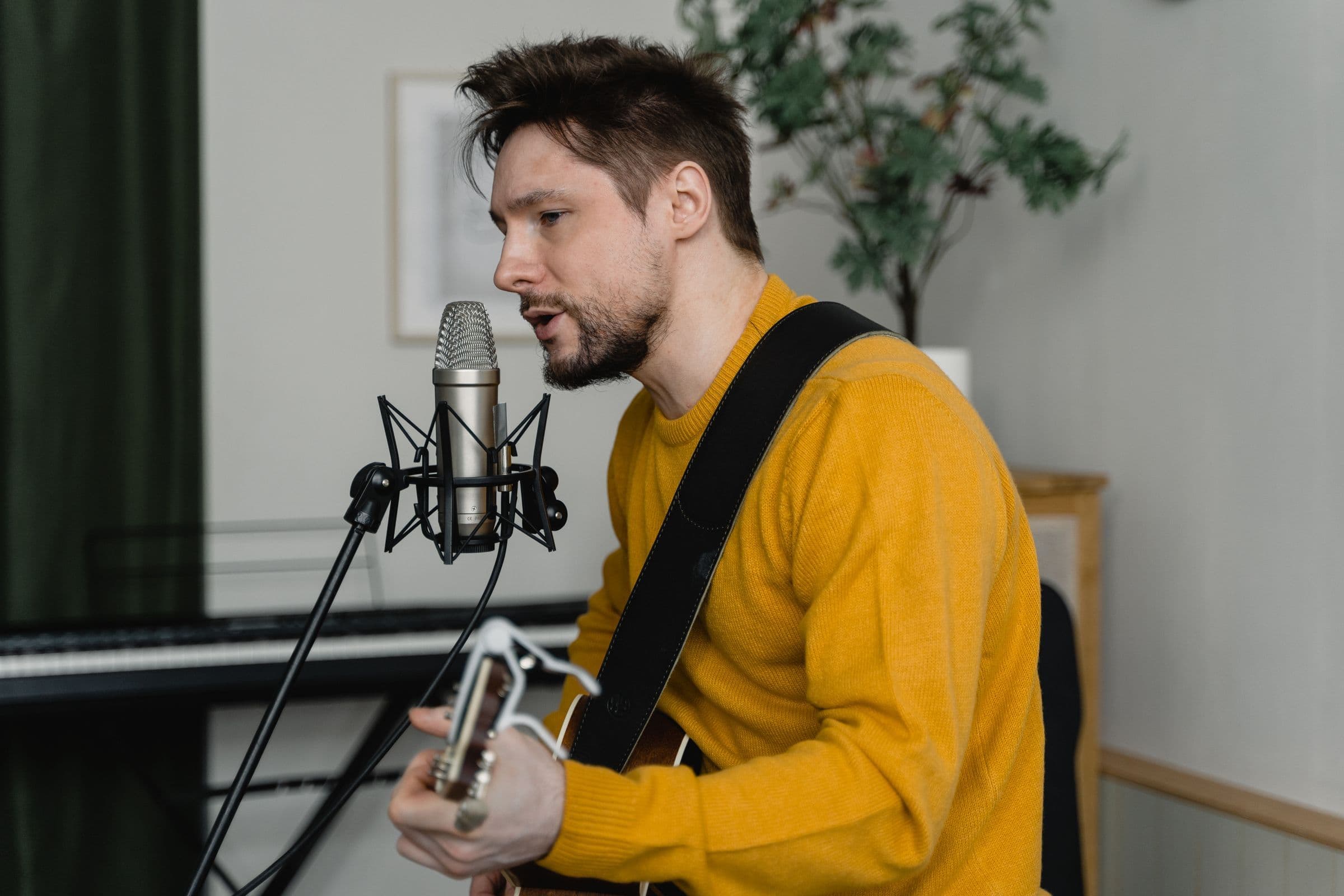 Man in a yellow sweater singing into a studio microphone while playing guitar