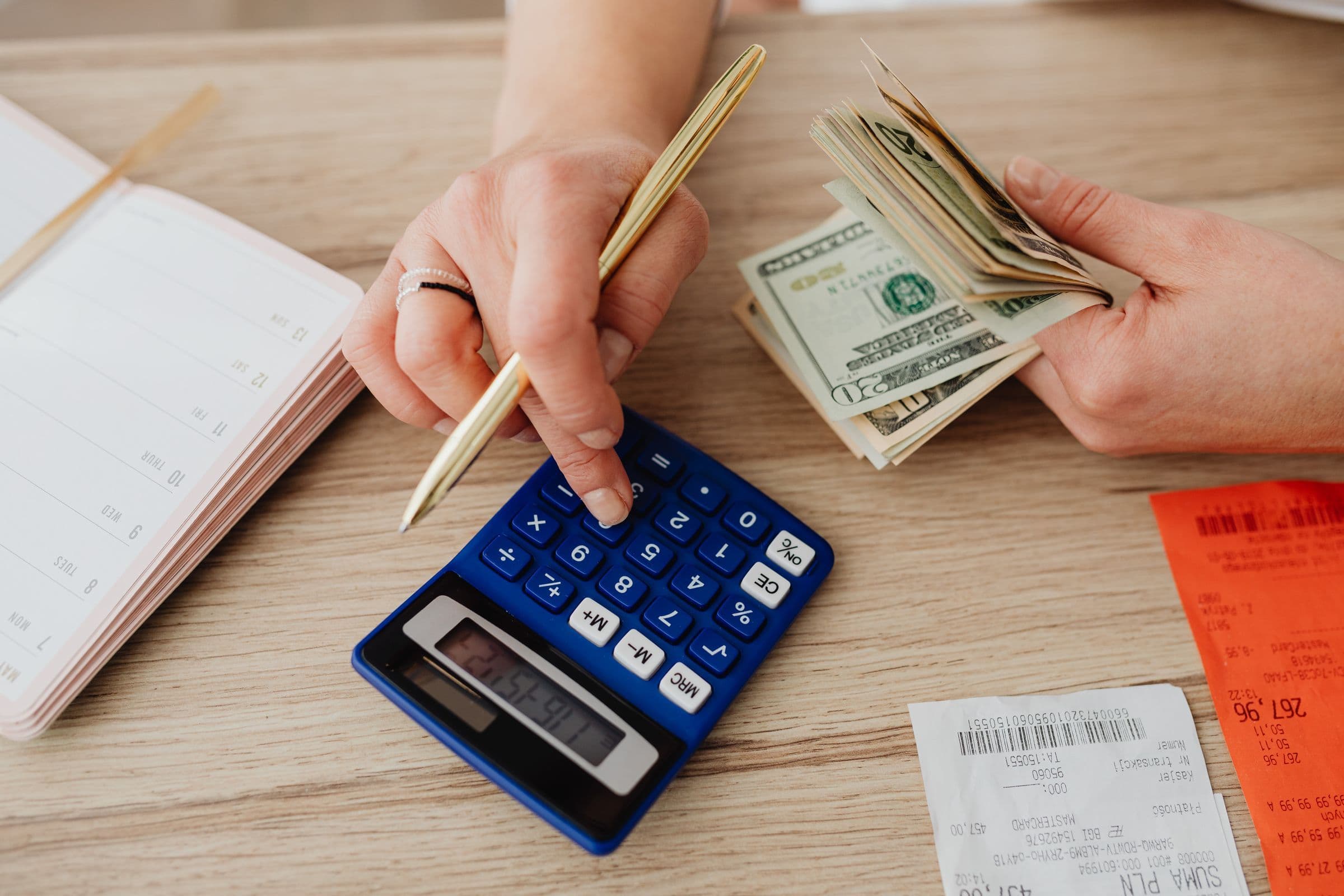 Hands holding dollar bills and a pen over a calculator, next to receipts and a notebook