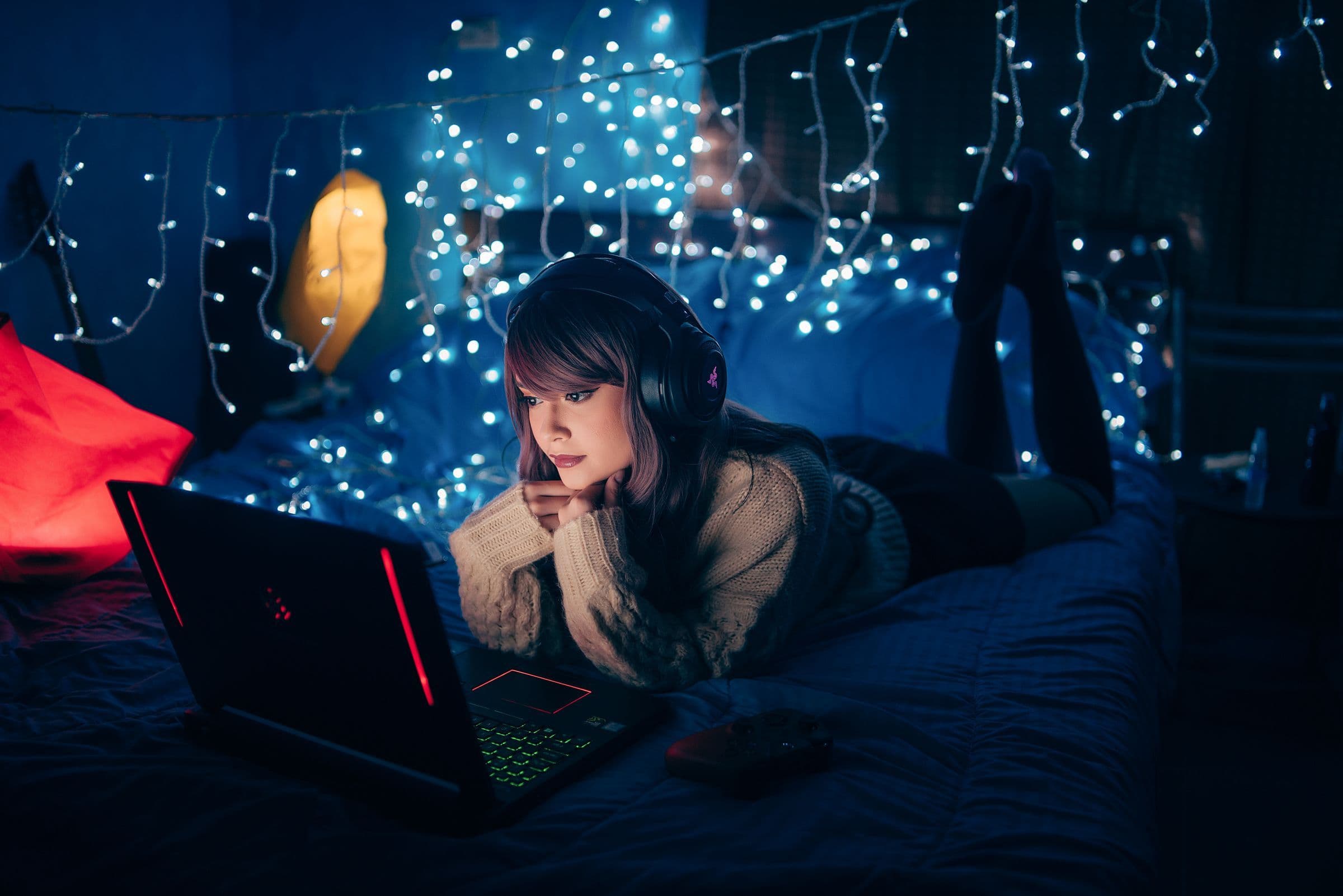 Woman in headphones lying on a bed at night, looking at a laptop under fairy lights