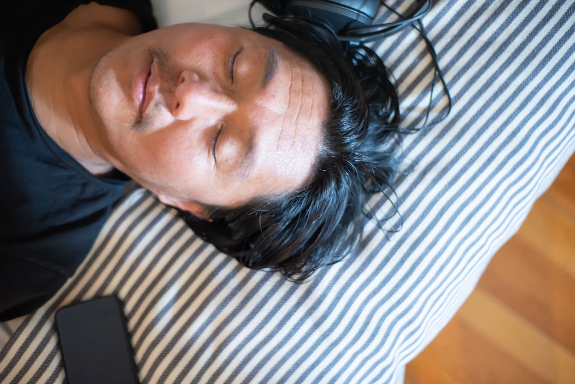 Man lying down with his eyes closed and headphones on, a phone beside him