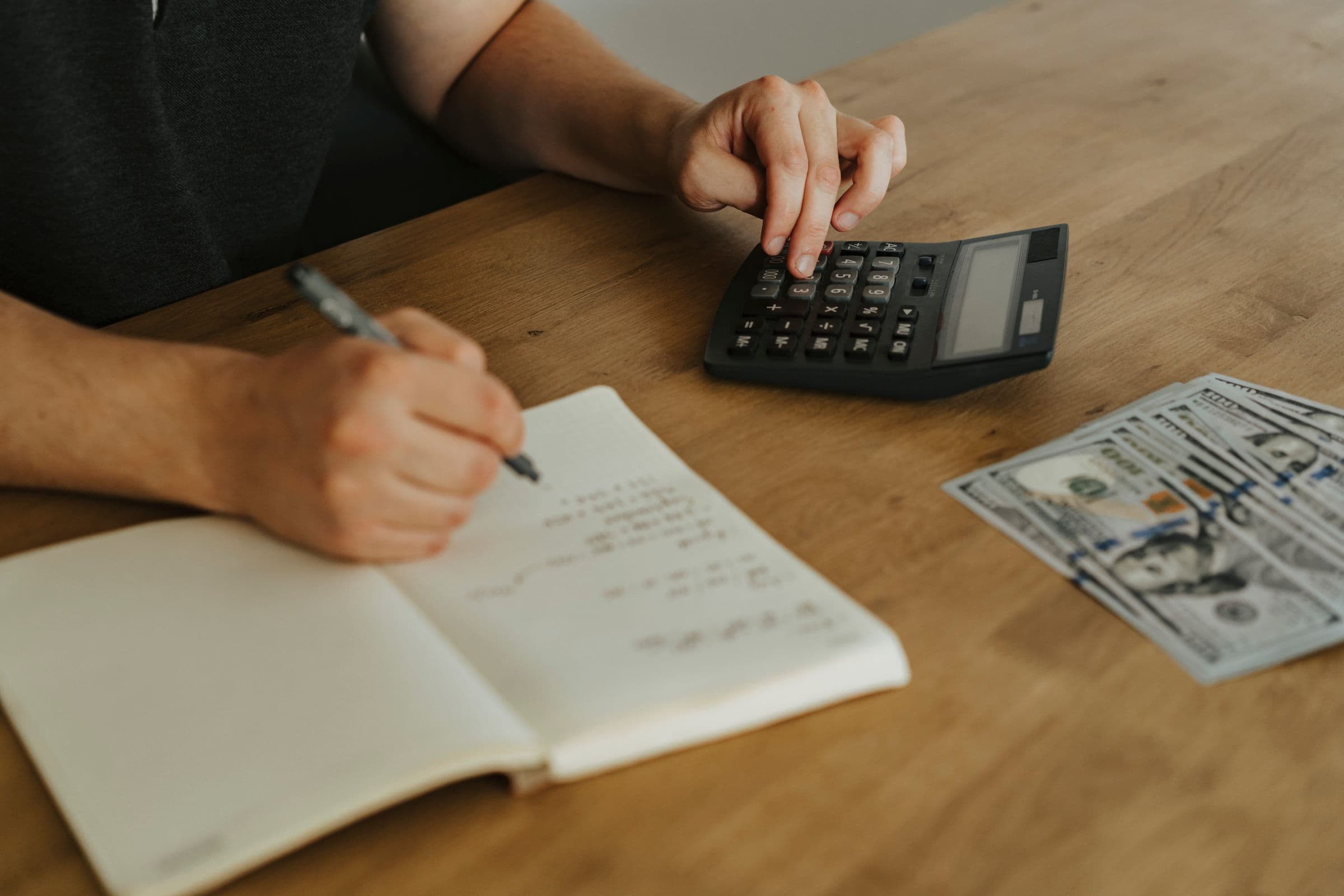 Hands working out a budget with a calculator, a notebook, and dollar bills on a wooden table