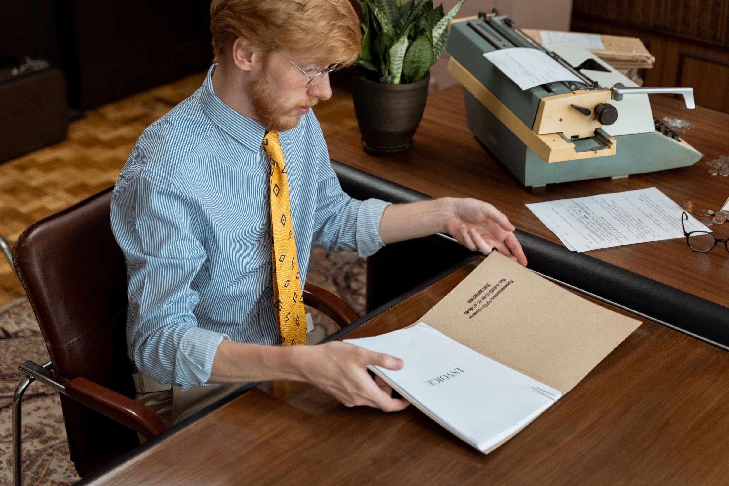 Man in a shirt and tie reading through a document at a wooden desk with a typewriter